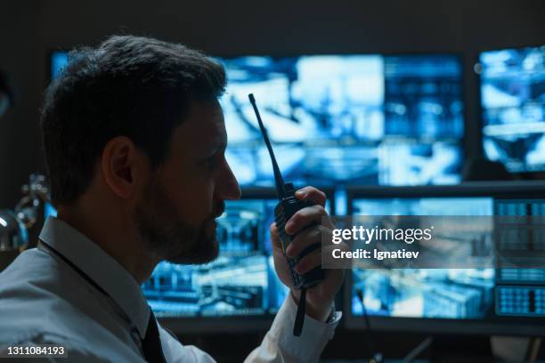 un homme à la barbe, un agent de sécurité ou un policier, peut-être des services de secours, parle sur un talkie-walkie sur fond de moniteurs numériques qui affichent des informations provenant des caméras de surveillance. - security guard photos et images de collection