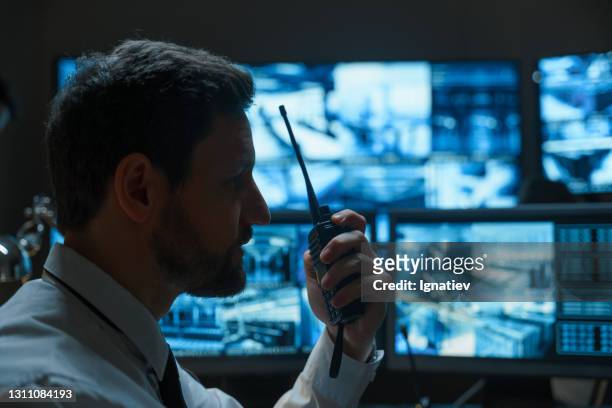 a man with a beard, a security guard or a police officer, possibly from the rescue service, is talking on a walkie-talkie against the background of digital monitors that display information from surveillance cameras. - security guard imagens e fotografias de stock
