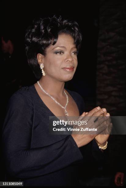 American talk show host and actress Oprah Winfrey, wearing a black outfit with a scoop neck, her hands raised with the fingertips touching, attends...