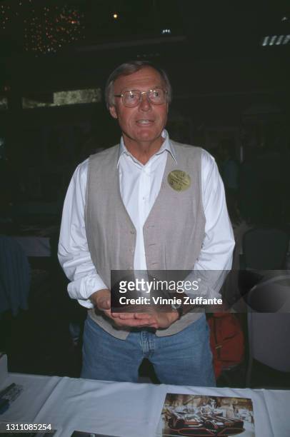 American actor Adam West , wearing a beige waistcoat over a white shirt, with blue jeans, on the table a photograph of the Batmobile from the...