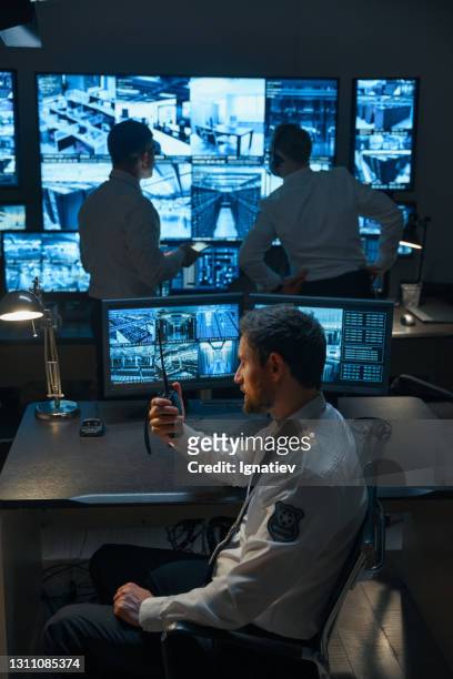 a man with a beard, a security guard or a police officer, possibly from the rescue service, is talking on a walkie-talkie against the background of digital monitors that display information from surveillance cameras. in the background, two of his colleagu - voice search stock pictures, royalty-free photos & images