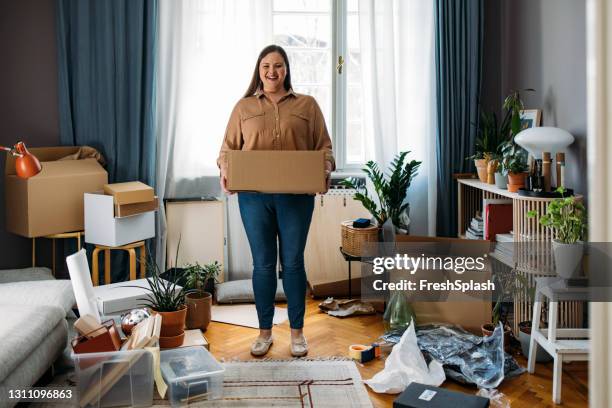 ready to move out: caucasian plus size woman standing in the middle of a messy room while holding a cardboard box and looking at camera ( wide angle horizontal shot) - messy living room stock pictures, royalty-free photos & images