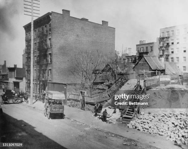 Carts parked along the sidewalk with a small wooden shed atop a mound of earth, reached by a flight of wooden steps in this image of Hell's Kitchen...