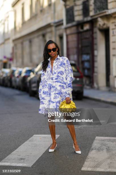 Emilie Joseph @in_fashionwetrust wears sunglasses, a blue and white Toile de Jouy shirt short dress from Moschino, a yellow shiny lime satin pouch...