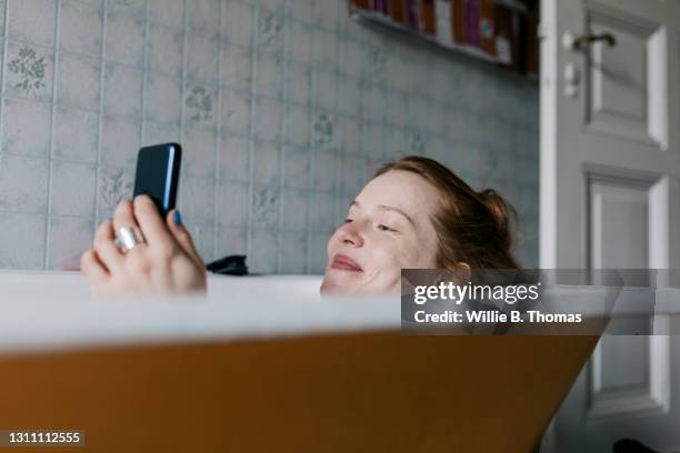 woman taking bath and smiling while messaging someone - buscador fotografías e imágenes de stock