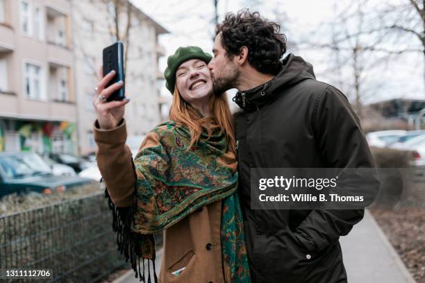 man kissing girlfriend on cheek while she takes selfie - couple smartphone happy stock pictures, royalty-free photos & images