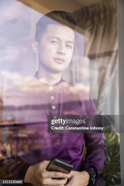 young man wearing baju melayu malay traditional costume behind window glass - baju melayu stock pictures, royalty-free photos & images