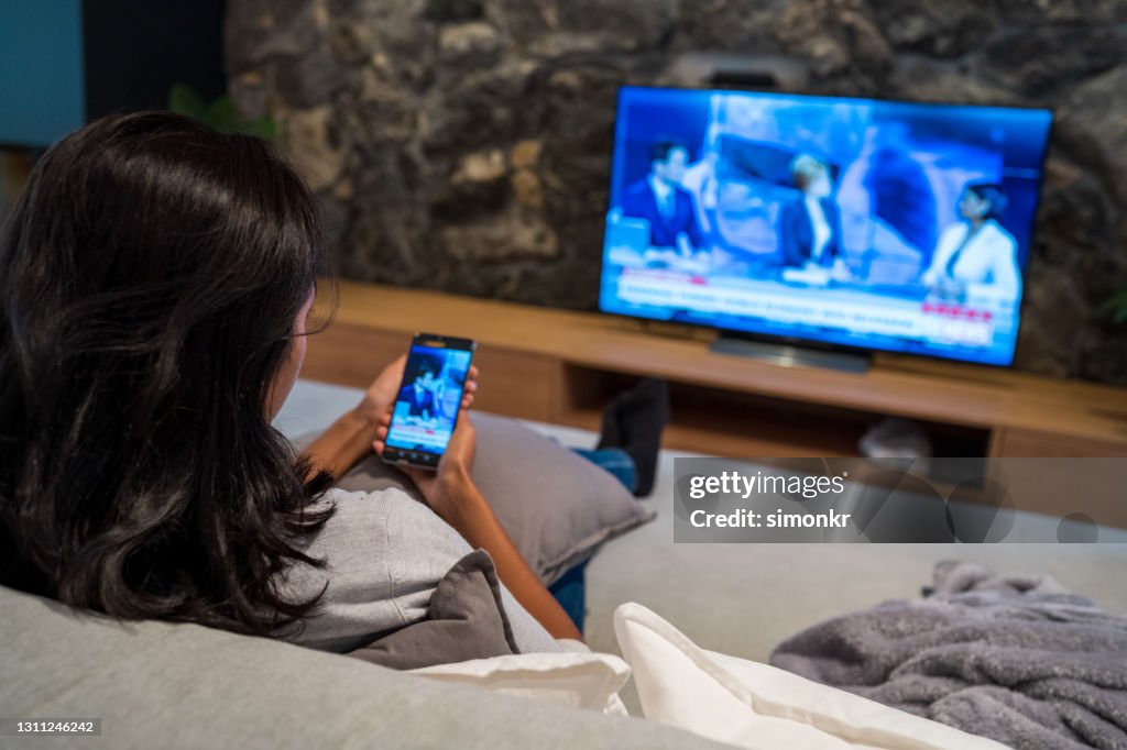 Jonge vrouw die nieuws op televisie en slimme telefoon bekijkt