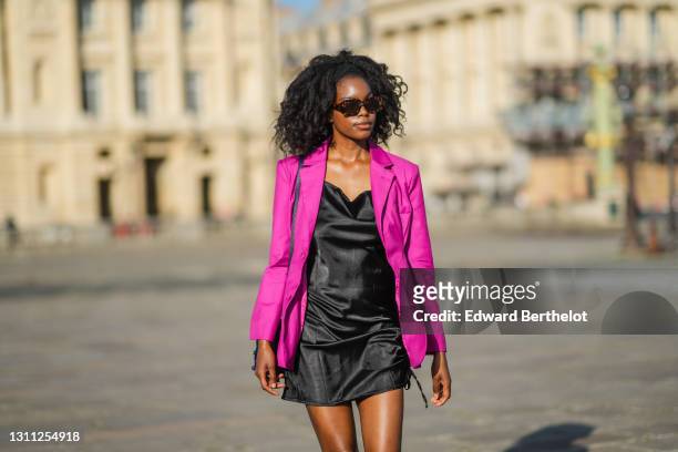 Magalie Kab wears sunglasses, a neon purple blazer jacket, a black short shiny satin / silky dress, a blue bejeweled bag, on March 31, 2021 in Paris,...
