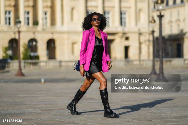 Magalie Kab wears sunglasses, a neon purple blazer jacket, a black short shiny satin / silky dress, a blue bejeweled bag, knee high black leather...