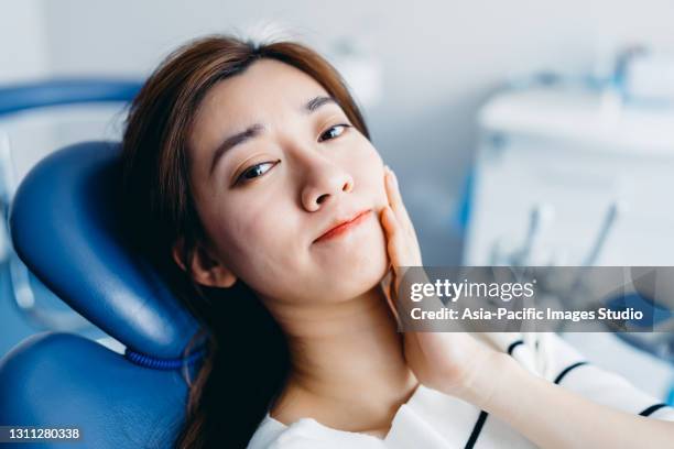 asiatische junge frau, die schmerzen verspürt und ihre wange mit der hand beim zahnarzt hält. zahnschmerzen konzept. - sensitivity in tooth stock-fotos und bilder