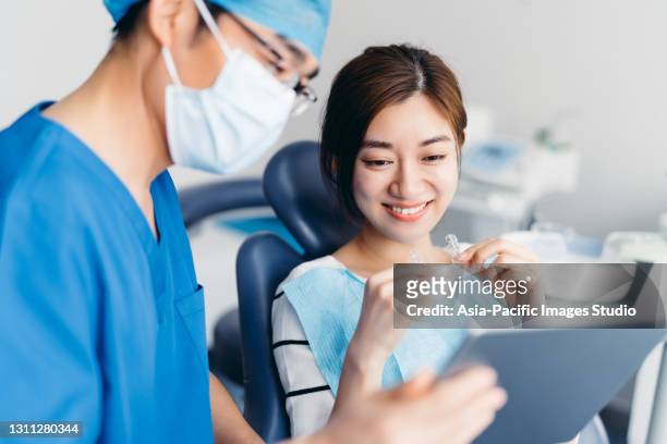 dentiste asiatique expliquant des rayons x de dent à un patient avec le comprimé numérique. jeune femme attirante asiatique retenant des mandats orthodontiques dans la clinique dentaire. concept d’orthodontie invisalign. - dentist office photos et images de collection