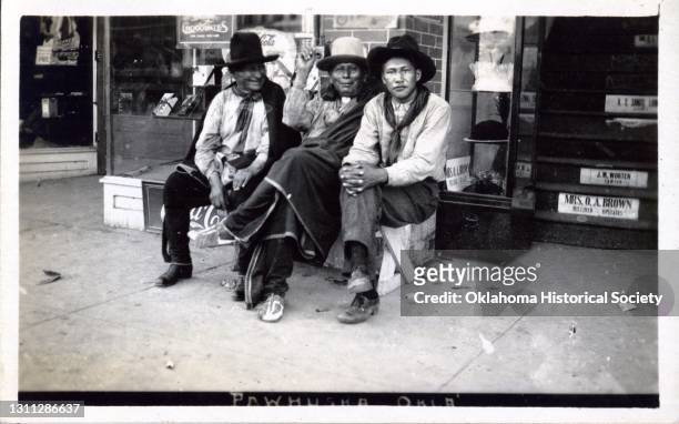 Postcard features a photograph of three of unidentified men of the Osage Nation as they sit on a bench in front of shop, Pawhuska, Oklahoma...