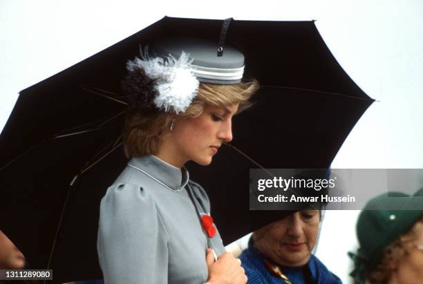 Diana, Princess of Wales, wearing a grey belted dress and a matching bell-boy style hat with feathers, holds an umbrella as she attends an Anzac Day...