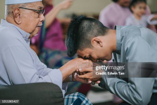 malay muslimischen enkel in traditioneller tracht zeigt entschuldigung geste an seinen großvater während aidilfitri feier malay familie zu hause feiern hari raya - hari raya celebration stock-fotos und bilder