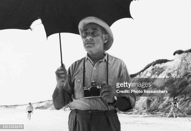 Portrait of German-born American photographer Alfred Eisenstaedt as he holds an umbrella and a camera on a beach, circa 1960s.