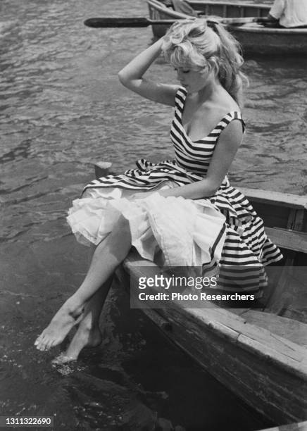 View of French actress Brigitte Bardot, in a striped dress, as she sits in the prow of a row boat and dips her feet in the water, 1950s.