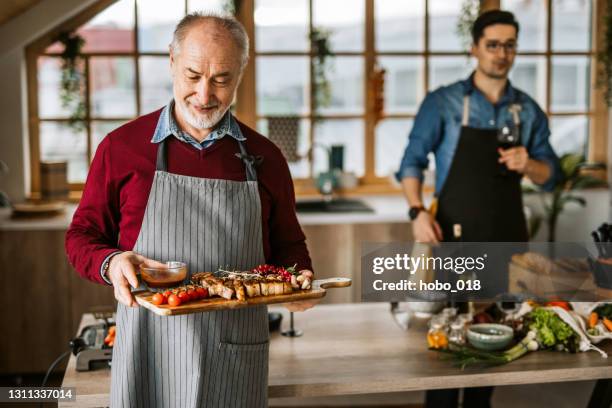 senior man in kitchen holding wooden board with served beef steak for dinner - serving dish stock pictures, royalty-free photos & images