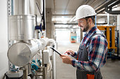Heating engineer worker holding tablet computer and setting parameters of heating system in factory boiler room.