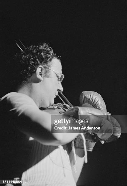 Humoriste français Coluche, de son vrai nom Michel Colucci, jouant du violon avec des gants de boxe, sur scène au théâtre Bobino à Paris.
