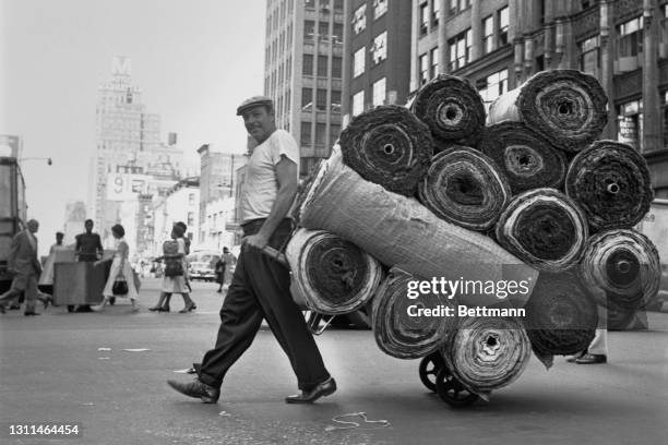 Deliveryman crossing Eighth Avenue with a sack truck loaded with rolls of fabric in the Garment District of New York City, New York, 29th August 1959.