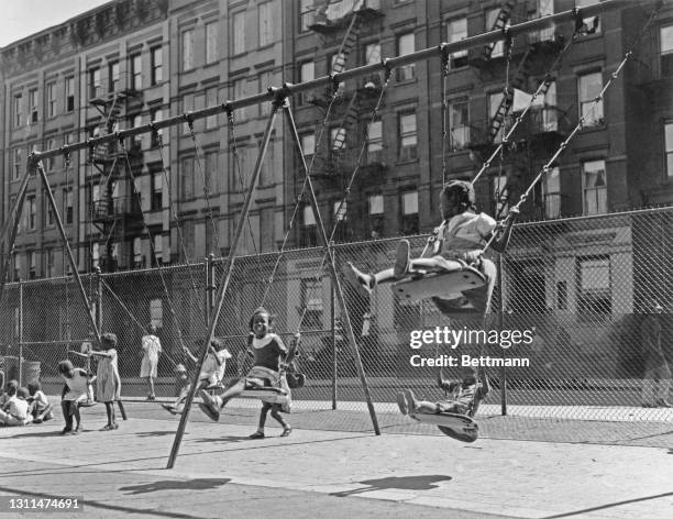 African-American children playing swings at a playground, with a chainlink fence in the background, and tenement blocks beyond that, in Harlem, New...