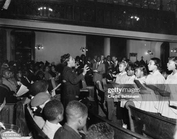 Choir dressed in surplices singing from their hymn books as they face the congregation, from which a woman stands to join the singing, during a...