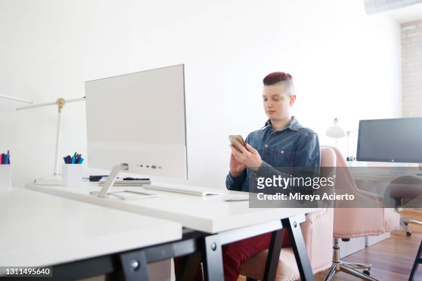A transgender millennial with red dyed hair, works at their desk while using the computer in a brightly lit office space wearing business casual clothing.
