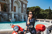 Beautiful young adult woman sitting on a motor scooter near Fontana dell'Acqua Paola in Rome, Italy