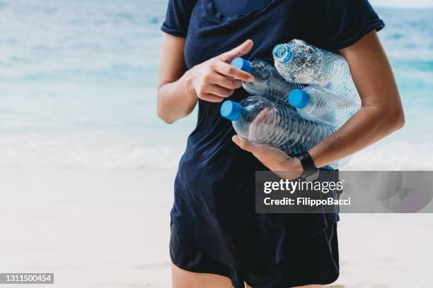 young adult woman picking up some abandoned plastic bottles on the beach - plastic bottle stock pictures, royalty-free photos & images