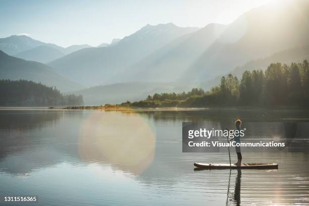 woman paddleboarding on calm lake in whistler during sunrise. - paddleboarding stock pictures, royalty-free photos & images