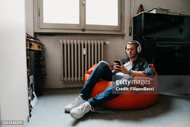joven adulto enviando mensajes de texto y escuchando música en su dormitorio - bean bags fotografías e imágenes de stock