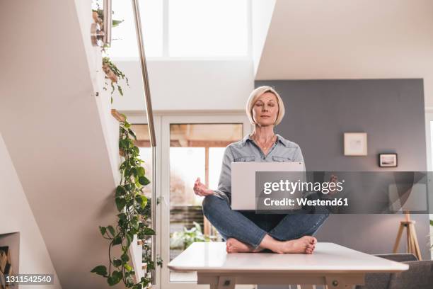 female entrepreneur with laptop sitting in lotus position on table at home - connected mindfulness work stock pictures, royalty-free photos & images