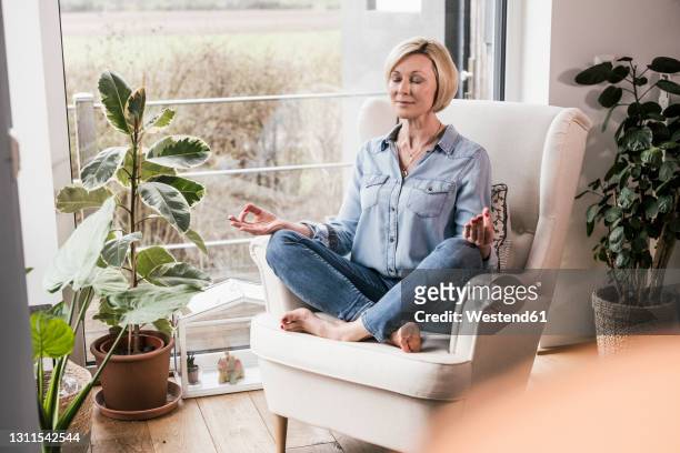 mature woman meditating while sitting on armchair in living room - yoga chair stockfoto's en -beelden