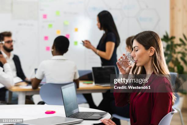 young businesswoman drinking water while sitting by laptop in office - wasser trinken büro stock-fotos und bilder