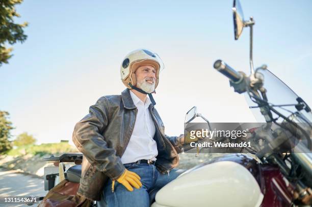 smiling man looking away while sitting on motorcycle - biker jacket stock pictures, royalty-free photos & images