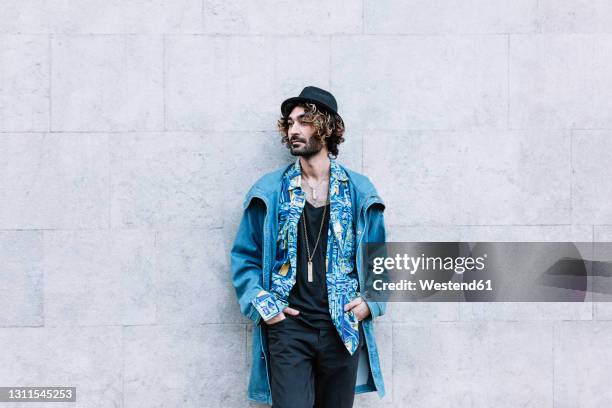 hipster man with hands in pockets looking away while standing in front of wall - jean jacket stock-fotos und bilder