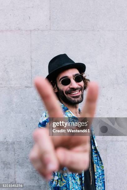 smiling young man showing peace sign while standing in front of wall - victory sign man stockfoto's en -beelden