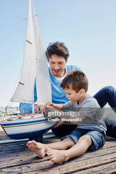 boy tying rope of toy boat while sitting with man on pier - father son sailing stock-fotos und bilder