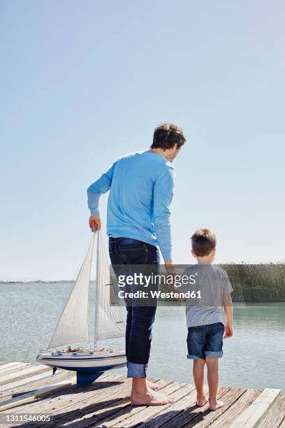 father with toy sailboat holding hand of son while standing on pier - father son sailing stock-fotos und bilder