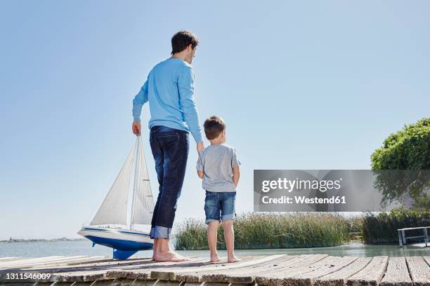 mature man with toy sailboat holding hand of boy while standing against sky - father son sailing stock pictures, royalty-free photos & images