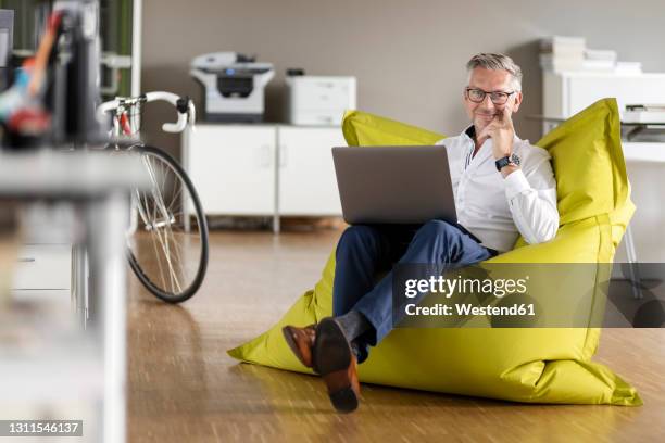 smiling businessman with laptop sitting on beanbag at open plan office - bean bags fotografías e imágenes de stock