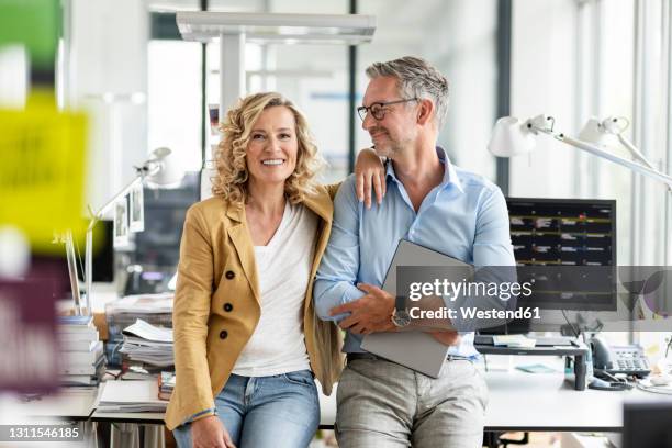 smiling businessman holding digital tablet while looking at female colleague while leaning at desk in office - 50 years in business stock pictures, royalty-free photos & images