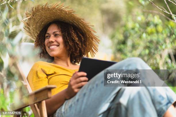 smiling woman in straw hat holding digital tablet while sitting on chair in vegetable garden - straw hat stock pictures, royalty-free photos & images