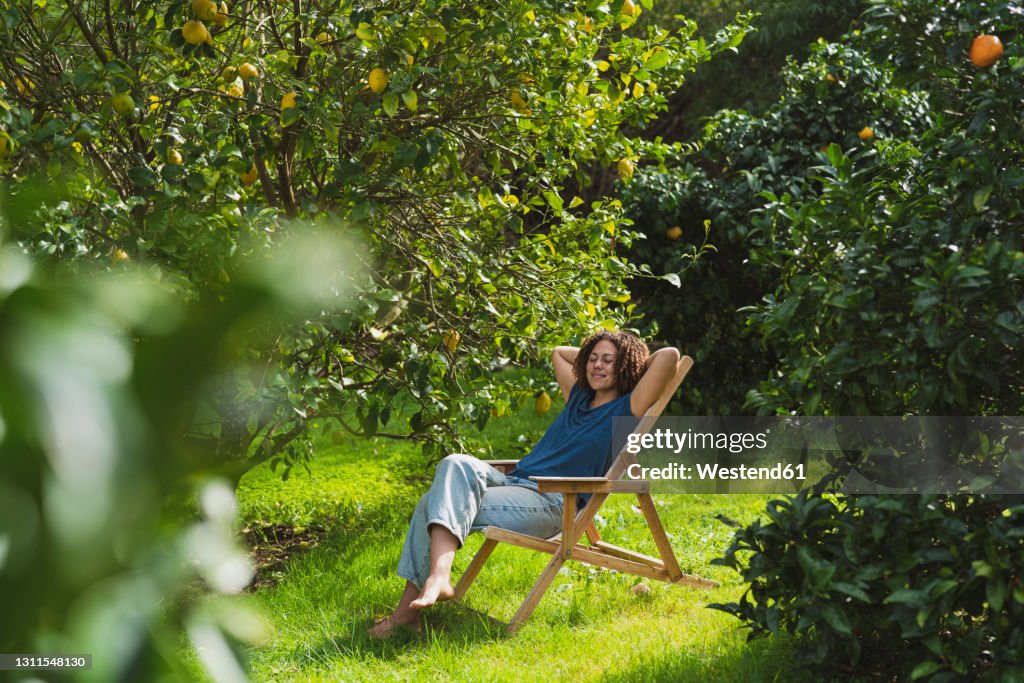 Woman with hands behind head relaxing on chair amidst trees in garden
