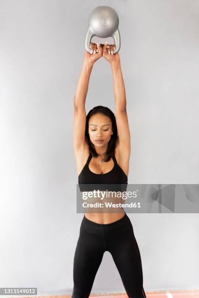 active young woman exercising with kettlebell in front of gray wall - kettle bells fotografías e imágenes de stock