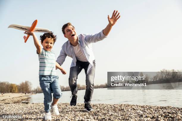 playful man and boy holding airplane toy while running by lakeshore on sunny day - airplane exterior stock pictures, royalty-free photos & images
