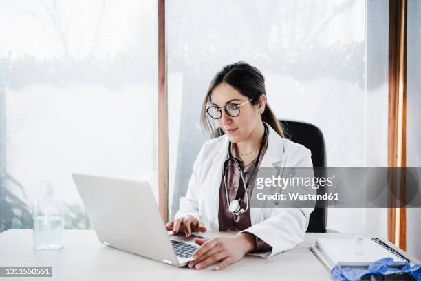 female medical expertise using laptop while sitting against window in hospital - doctor on computer stockfoto's en -beelden