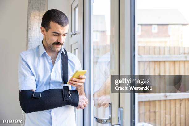 man wearing arm sling using smart phone while standing by window at home - broken arm stock pictures, royalty-free photos & images