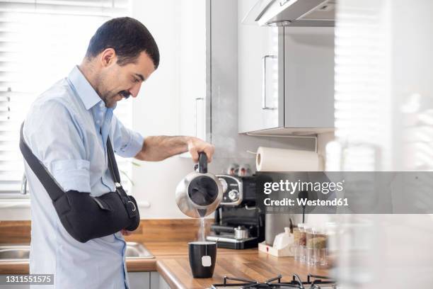 injured man with arm sling pouring tea in cup through kettle in kitchen at home - gebrochener arm stock-fotos und bilder